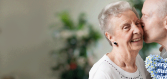 Pensionist küsst Pensionistin auf die Wange, welche fröhlich lächelt. Darunter sieht man ein Herz aus Vergissmeinnicht Blumen mit der gleichnamigen Aufschrift auf dem Herz stehend