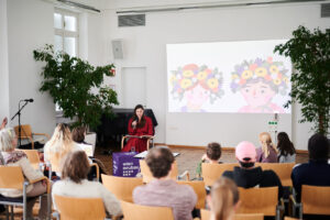 Buchvorlesung, Autorin liest aus dem Buch, während Illustrationen an einer Leinwand projiziert werden