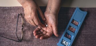 ältere Hände mit Tabletten in der Hand liegen auf einem Tisch, daneben liegt eine Dosierungsdose und eine Brille.