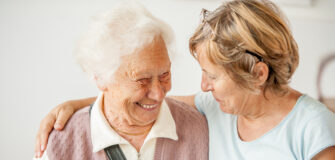 Eine Person mittleren Alters legt den Arm um eine ältere Person und sieht sie lächelnd an. Die ältere Person lächelt.